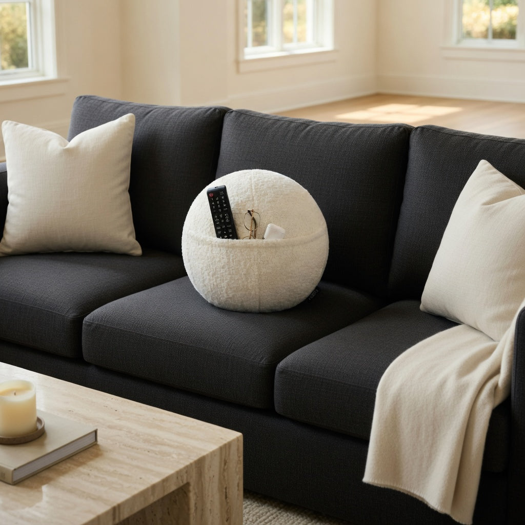 The COZELA Orb bouclé sphere pillow sitting on a dark gray sectional sofa with a remote, glasses, and AirPods case tucked into its hidden pocket