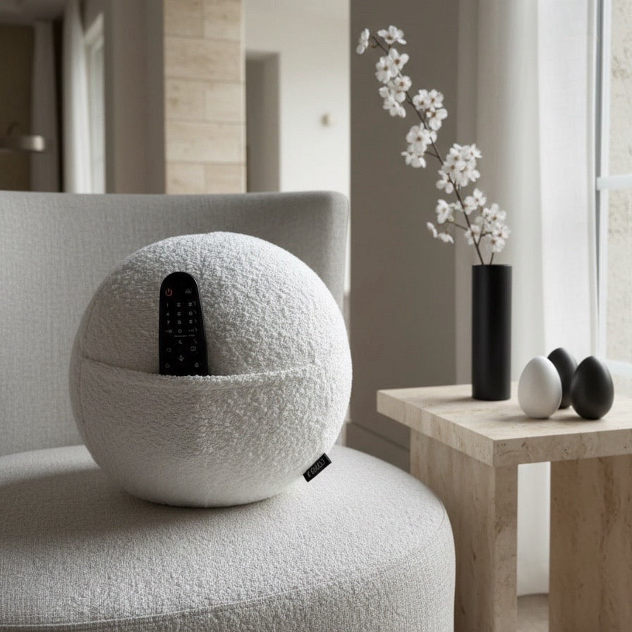 COZELA Orb bouclé sphere pillow with a remote in the hidden pocket, styled on a chair beside a travertine side table with cherry blossoms